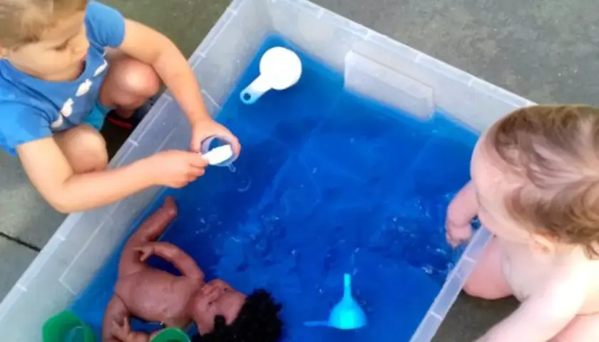 Niños de 3 a 4 años disfrutan de juegos acuáticos en el colegio, explorando con una muñeca y recipientes.