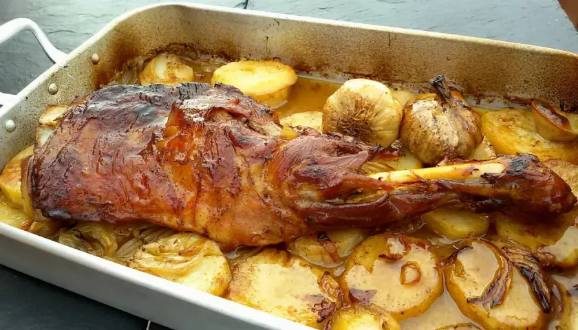 Paletilla de cordero al horno dorada y jugosa, acompañada de patatas asadas y ajos tiernos en una bandeja.
