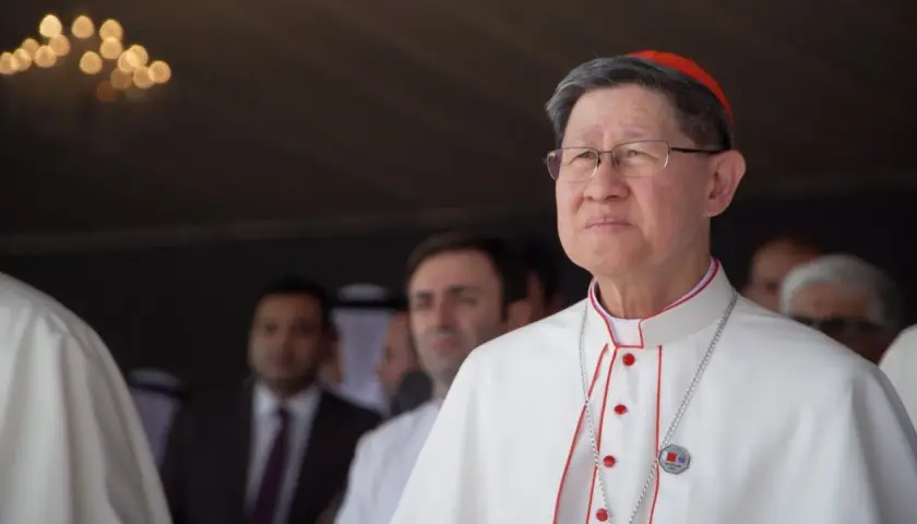 Kardinal Tagle, mit Brille und roter Pileolus, trägt ein weißes Gewand mit roten Akzenten. Im Hintergrund unscharfe Männer.