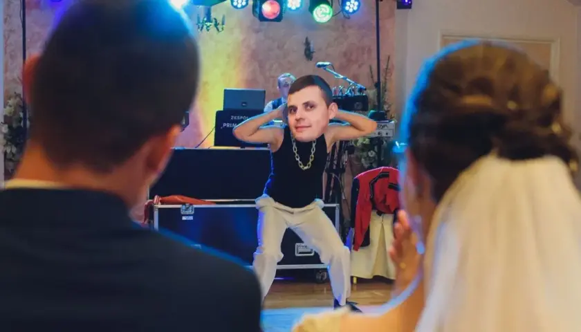 Zabawny moment podczas oczepin weselnych. Pan młody tańczy z wielką głową na szyi, a para młoda obserwuje go z uśmiechem.