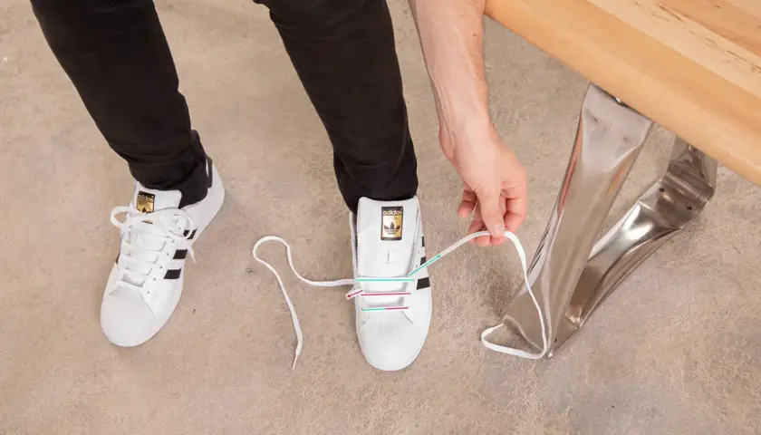 Instrukcja jak wiązać sneakersy: białe buty Adidas z czarnymi paskami, osoba zawiązuje sznurówki.