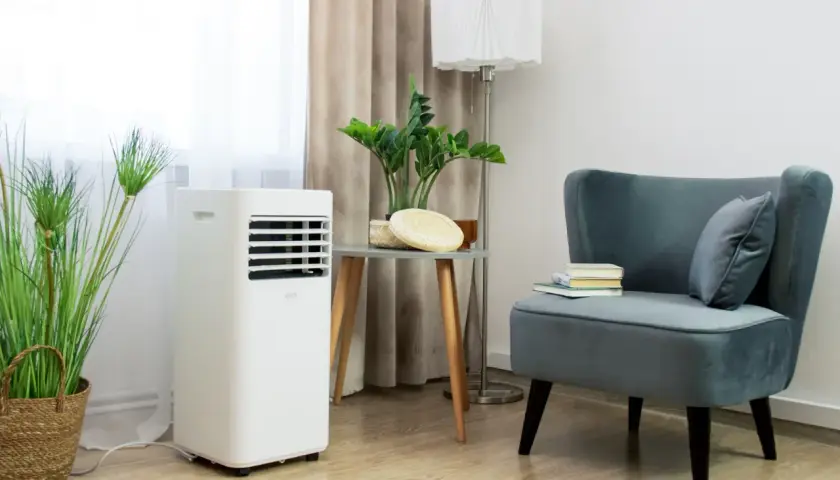 Biały klimatyzator przenośny stoi obok rośliny w wiklinowym koszu. Wygodny fotel z książkami i poduszką dopełnia wnętrze.