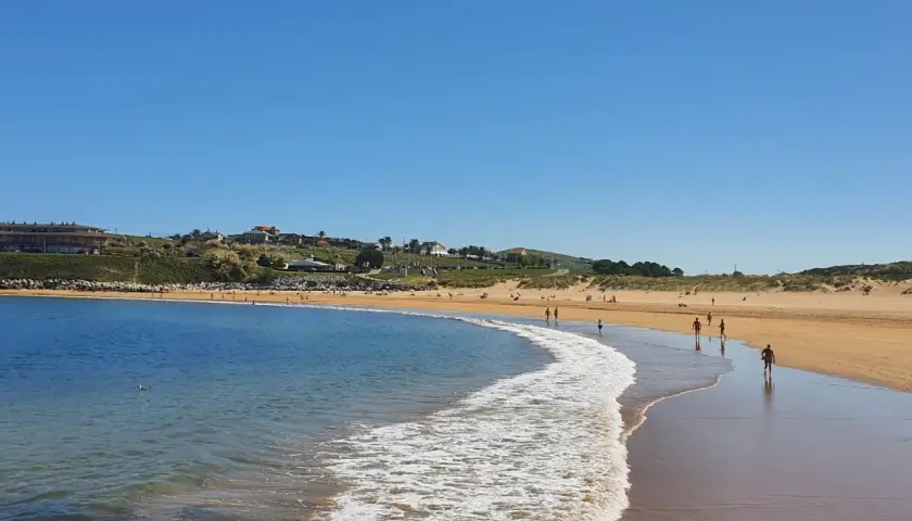 La **playa de Cuchía** se extiende bajo un cielo azul, con gente paseando por la arena dorada y el mar acariciando la costa.
