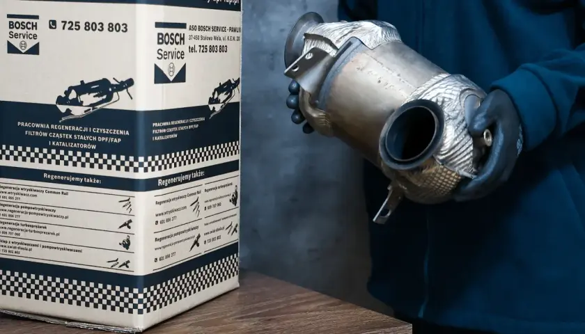 Osoba w rękawiczkach trzyma element układu wydechowego, prawdopodobnie filtr DPF. Obok stoi karton z logo Bosch Service, informujący o regeneracji filtrów DPF w dieslu.