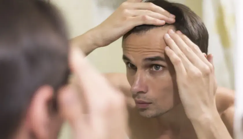 Hombre joven se mira al espejo, tocando su frente, preocupado por la pérdida de cabello.