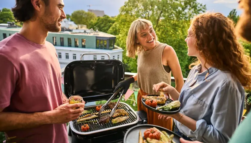 Vänner njuter av en grillfest på balkongen med en bärbar kolgrill. Saftiga biffar och grönsaker grillas medan solen går ner.