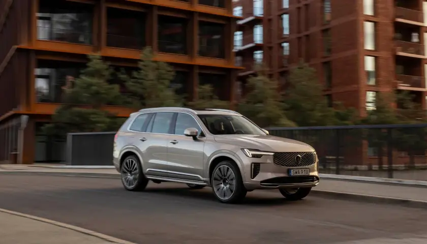 En ljusbeige Volvo SUV kör på en stadsgata. Bakgrunden visar moderna tegelbyggnader och gröna träd. Bilen visar upp olika volvo utrustningsnivåer.