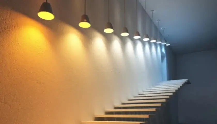 Escalera con luces colgantes que crean un degradado de luz y color, desde cálido hasta frío, iluminando la pared texturizada.