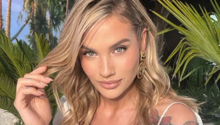 Karolina Gilon z blond włosami, niebieskimi oczami i tatuażem na ramieniu, na tle roślinności.