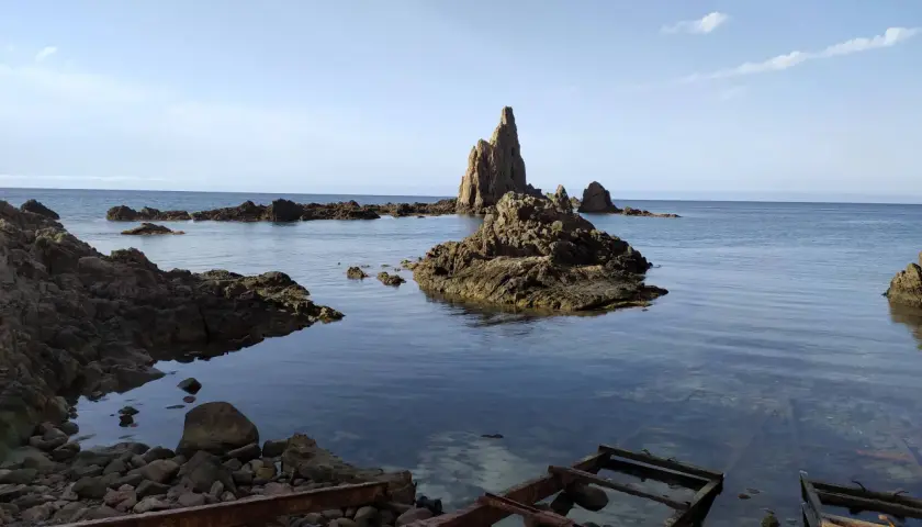 Rocas escarpadas emergen del mar azul, evocando el misterio del arrecife de las sirenas. El agua cristalina revela el lecho rocoso y restos oxidados de estructuras antiguas.