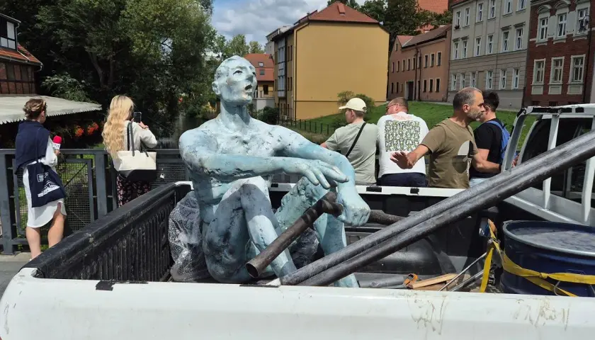 Rzeźbiarze transportują niebieską rzeźbę siedzącego mężczyzny w bagażniku pickupa. W tle widać budynki i ludzi.