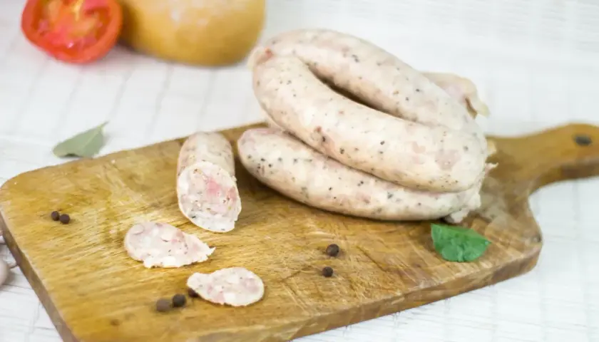 Biała kiełbasa na desce, obok pomidor, czosnek i przyprawy. Zastanawiasz się, ile czasu moczymy jelita do kiełbasy?