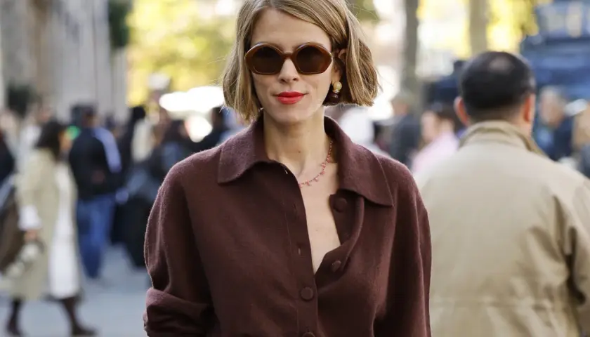 Mujer con corte francés con raya lateral, gafas oscuras y labial rojo, luciendo un suéter marrón.