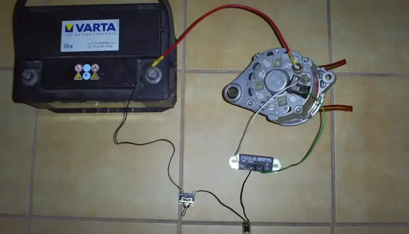 Akumulator Varta, alternator, regulator napięcia i żarówka połączone przewodami na podłodze.