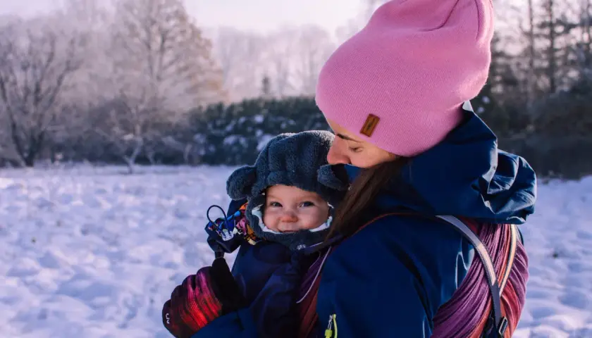 Mama w różowej czapce i granatowej kurtce tuli dziecko w nosidełku. Zima w pełni, śnieg wokół. Jak ubrać dziecko do nosidełka zimą? Warstwowo i ciepło!