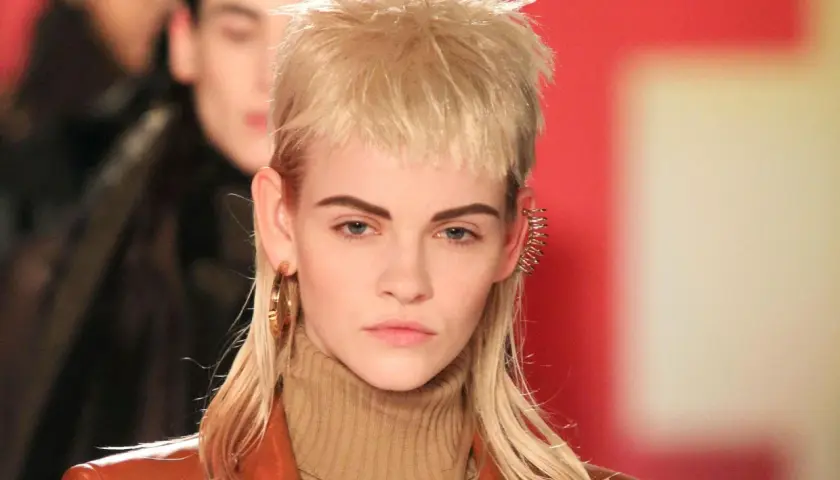 Ein Model präsentiert einen modernen Mullet mit blonden Locken, auffälligen Ohrringen und einem braunen Rollkragenpullover.