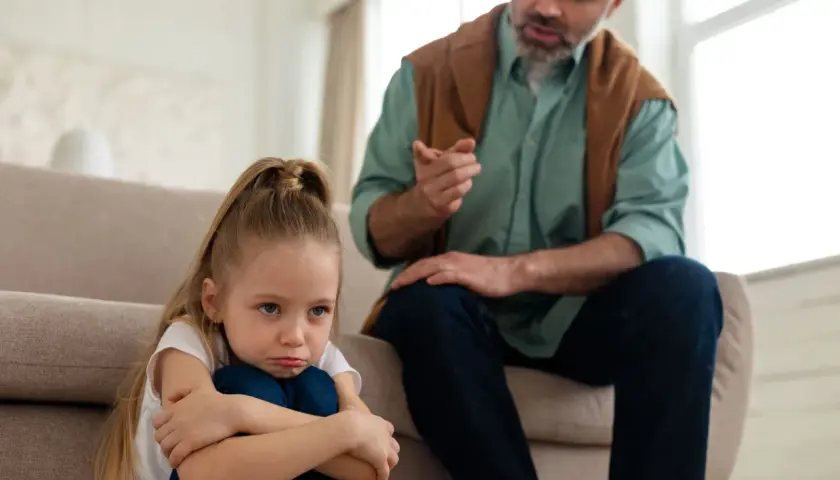 Smutna dziewczynka siedzi na podłodze, podczas gdy ojciec mówi do niej ostro, ukazując typowe daddy issues zachowania.