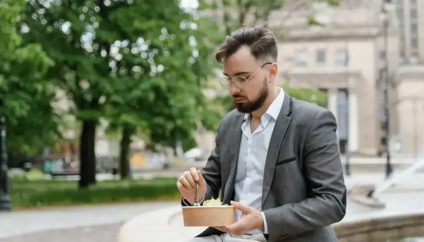 Mężczyzna w okularach i marynarce robi sobie przerwę w pracy, jedząc sałatkę na ławce w parku.