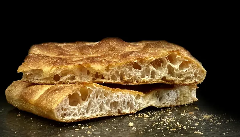 Schiacciata toscana dorata e fragrante, tagliata a metà, con alveolatura evidente e briciole sparse.