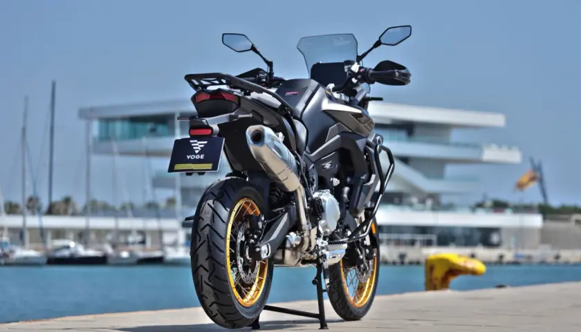 Voge 900 DSX ficha técnica: una moto de aventura negra con detalles dorados, aparcada junto a un puerto con veleros y un edificio moderno.