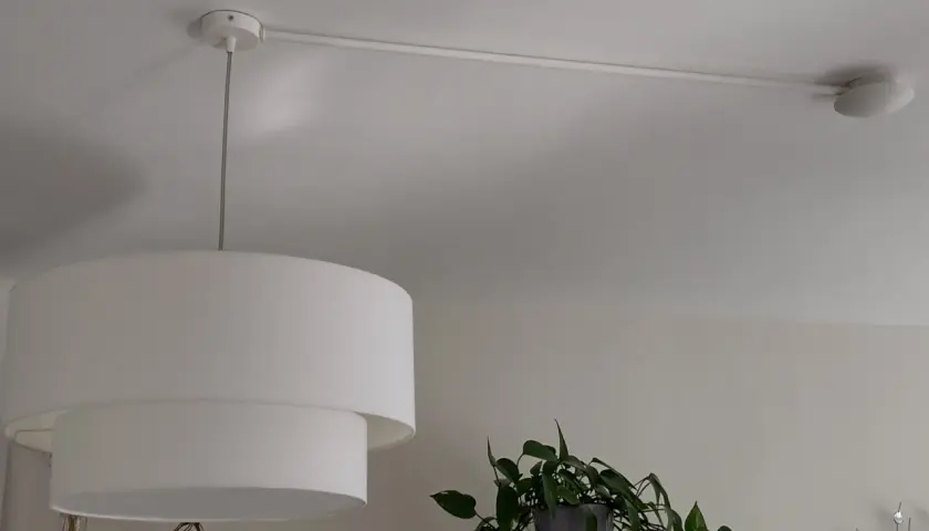Lámpara de techo con un punto de luz desplazado, colgada de un riel blanco. Una planta verde asoma por la derecha.