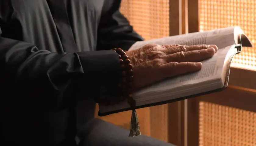 Dłoń starszego mężczyzny spoczywa na otwartej Biblii. Wokół nadgarstka ma drewniane paciorki. Gdzie jest pastor?