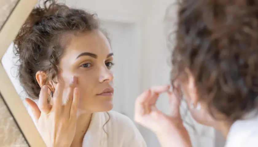 Woman applying cream to her face in a mirror.