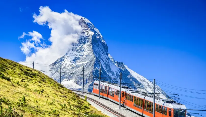 An orange cogwheel train ascends a grassy slope towards the majestic Matterhorn, a perfect scene for hiking in Switzerland.