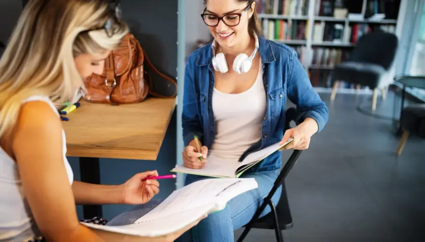 Dwie studentki uczą się do egzaminów w bibliotece. Jedna z nich, w okularach i dżinsowej koszuli, uśmiecha się, pisząc w zeszycie. To idealne miejsce do nauki w **liceum dla dorosłych 2-letnim**.