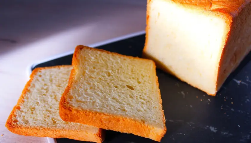 Pain de mie moelleux, parfait pour une recette pain de mie boulangerie maison. Deux tranches dorées et un pain entier sur un plateau sombre.