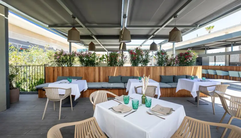 Terraza de la colonia de carmela con mesas puestas, asientos de madera y plantas floridas.