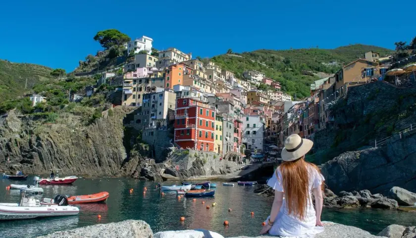 A woman in a white dress and hat gazes at the colorful houses of Riomaggiore, one of the best towns to stay in Cinque Terre, nestled on a cliffside overlooking the harbor.