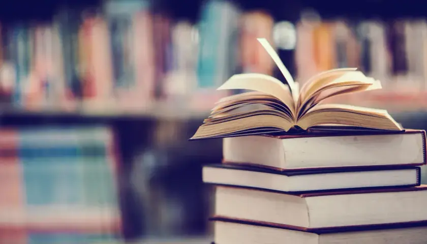 Stos książek, na wierzchu otwarta księga. W tle regały pełne literatury, w tym podręczniki do prawa pracy.