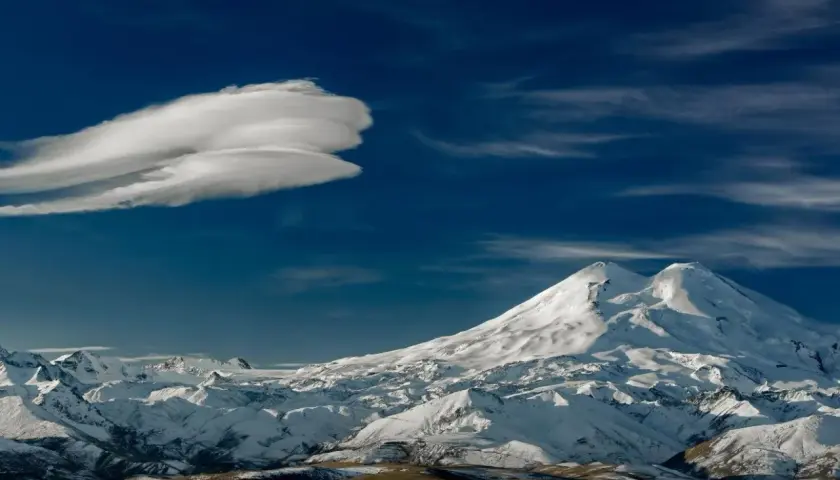Pokryty śniegiem Elbrus, najwyższy szczyt Rosji, góruje nad ośnieżonymi pasmami górskimi pod błękitnym niebem z charakterystyczną chmurą.