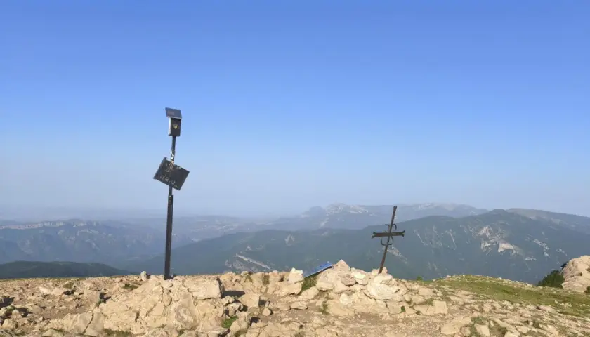 En el cap de la gallina pelada, un poste con placa y un sol solar vigilan el paisaje montañoso. A su lado, una cruz metálica se alza sobre rocas.