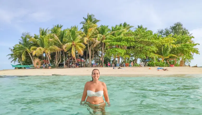 Mujer sonríe en aguas cristalinas, con una isla tropical llena de palmeras al fondo. ¡Un sueño para viajar a Filipinas!