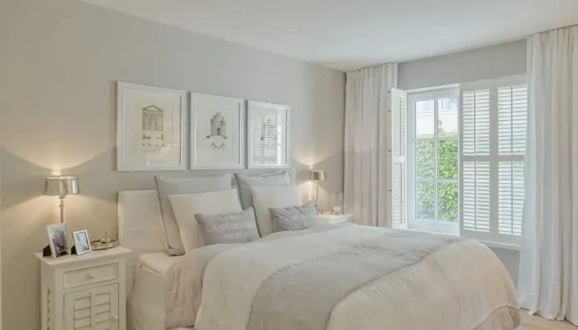 Helles, luftiges Schlafzimmer im Strandhaus-Stil mit weißem Bett, Kissen und Gardinen. Drei gerahmte Zeichnungen über dem Bett.
