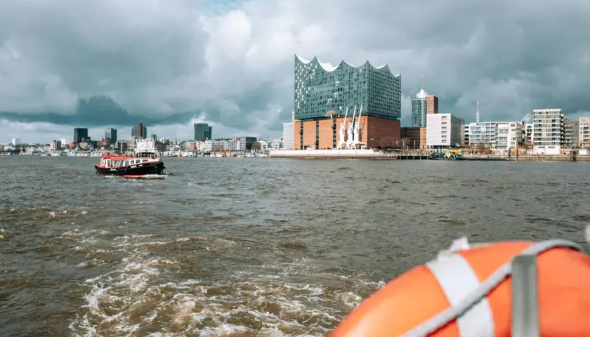 Blick vom Wasser auf Hamburgs Elbphilharmonie und die Skyline. Eine kleine Barkasse fährt vorbei. Welche Hafenrundfahrt in Hamburg ist die beste?