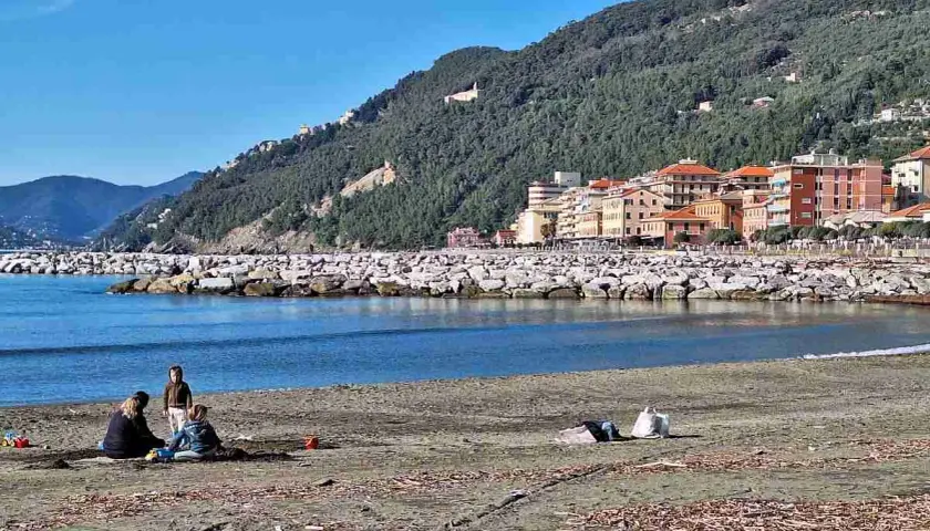 Vue de la plage de Chiavari avec des familles profitant du soleil, des bâtiments colorés et une colline verdoyante en arrière-plan.