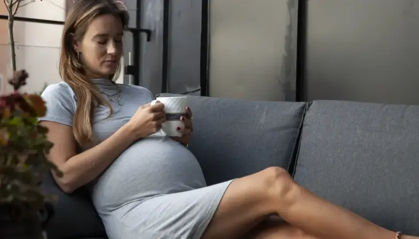 En gravid kvinna i sitt andra trimester vilar på en soffa med en kopp i handen.