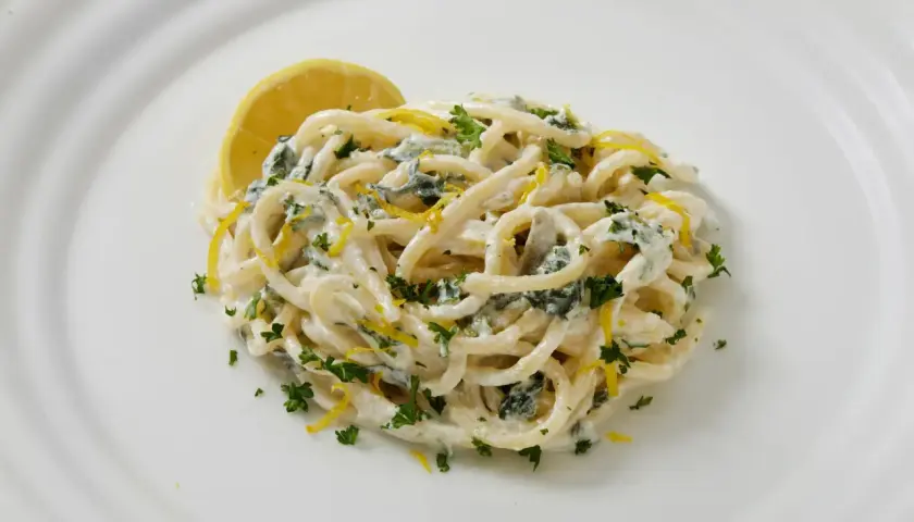 Un plat de pasta al limone crémeuse, garnie de persil frais et de zeste de citron, avec une tranche de citron sur le côté.