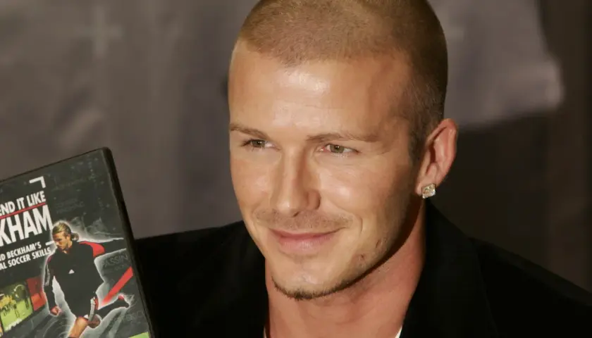 David Beckham, con su característico corte afeitado, sonríe mientras sostiene un DVD de sus habilidades de fútbol.