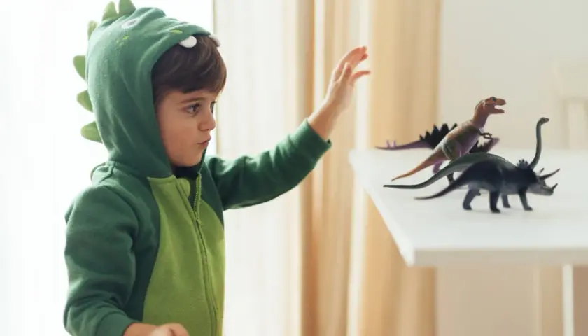 Chłopiec w zielonym stroju dinozaura DIY sięga po figurki prehistorycznych gadów.