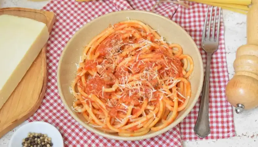 Miseczka pełna aromatycznego makaronu amatriciana, posypanego startym parmezanem. Obok kawałek sera, ziarna pieprzu i widelec.