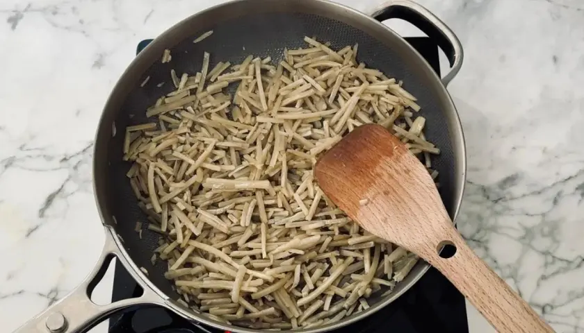 Strips of burdock root are being stir-fried in a pan, a key step in preparing a delicious kinpira recipe. A wooden spatula is used to toss the ingredients.