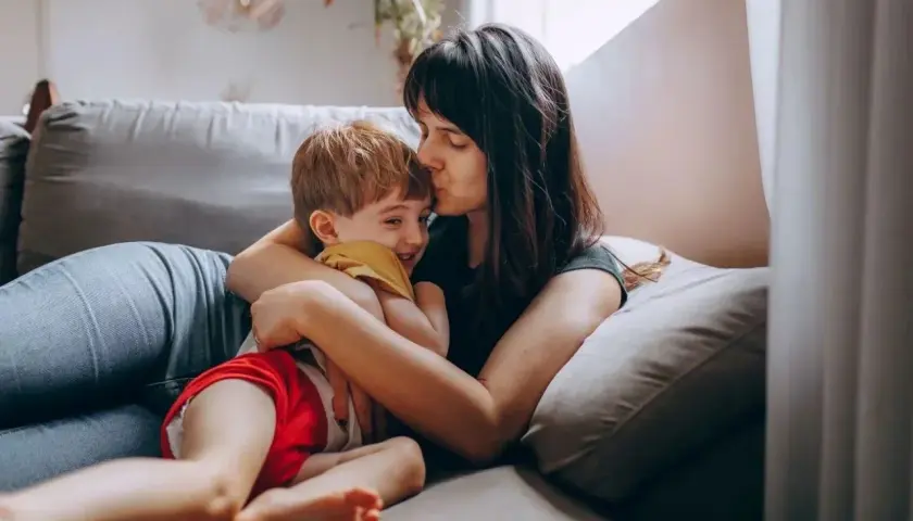 Mama całuje synka w czoło, przytulając go na kanapie. Dziecko śmieje się radośnie. Może dziś nie posyłam dziecka do przedszkola.