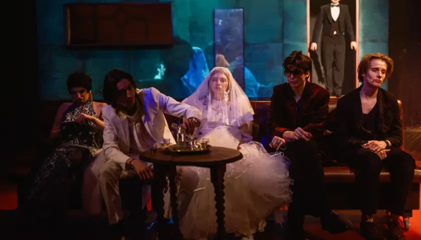 Scena z teatru AST Wrocław. Panna młoda w welonie, pan młody w garniturze i inni bohaterowie siedzą na kanapie. Sprawdź repertuar!