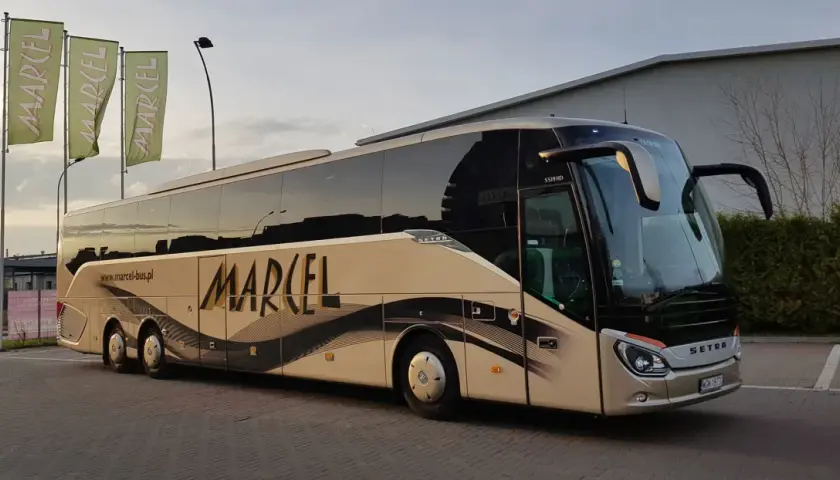Autobus Marcel z napisem na boku, stojący na parkingu, z zielonymi flagami w tle.