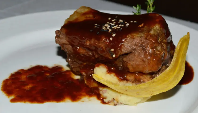 Un plat de ribs de boeuf barbecue, nappé d'une sauce foncée, servi avec une purée et une tranche de banane plantain frite.