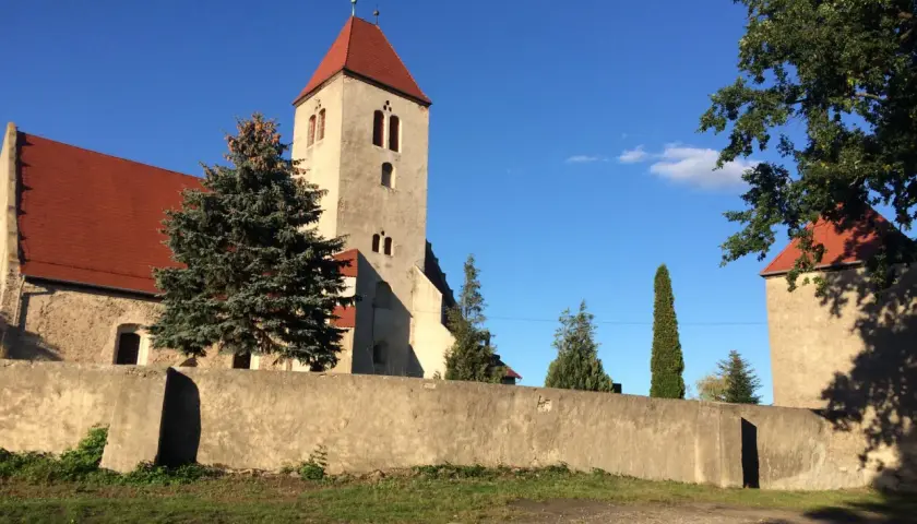 Kamienna wieża kościelna z czerwonym dachem w Nowogród Bobrzański, otoczona murem i zielenią.